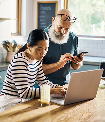 Couple using a laptop and phone in the kitchen at home to check account.