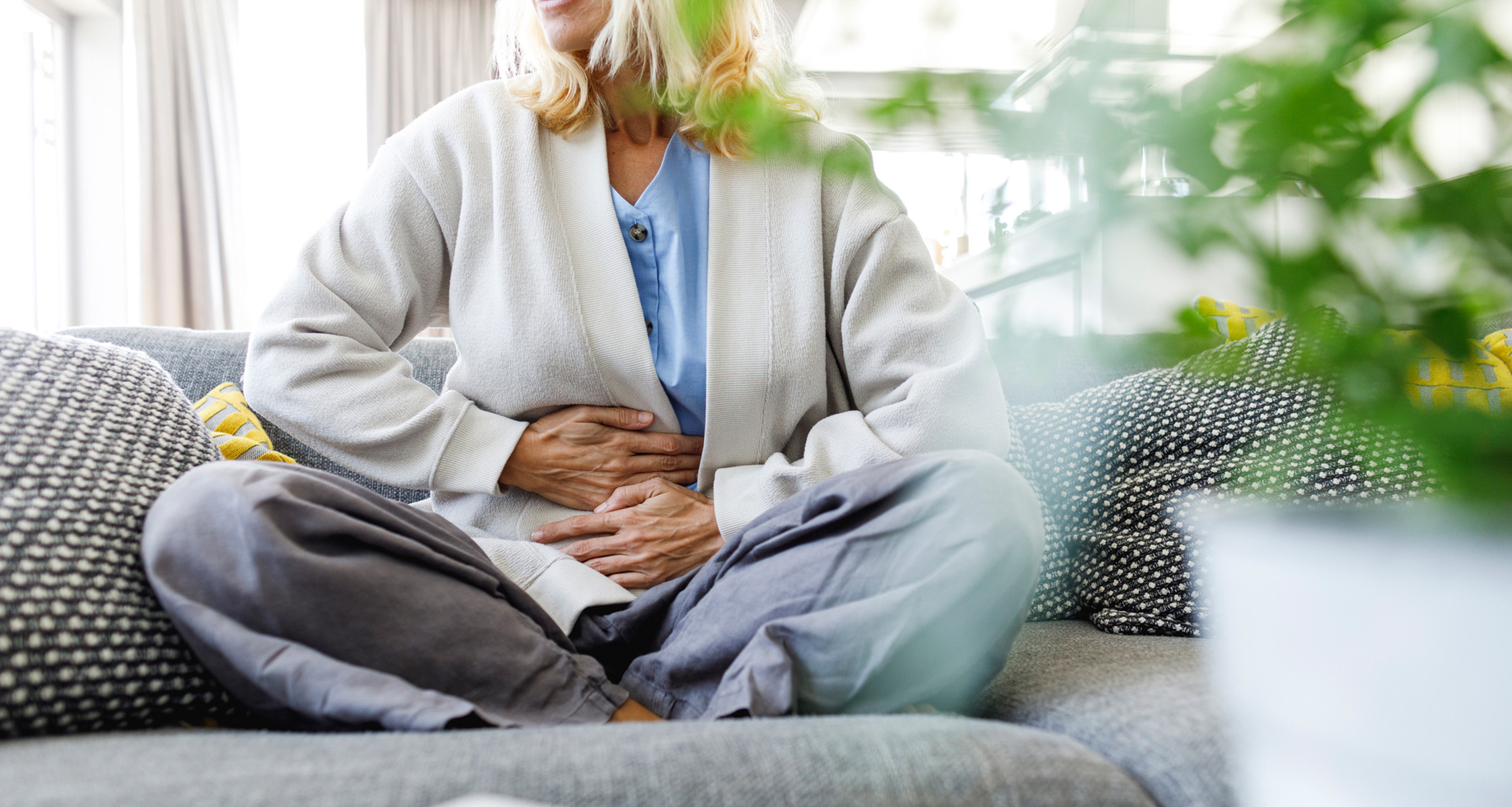 A woman sitting on a couch, indicating abdominal pain or discomfort, a common symptom of lupus.