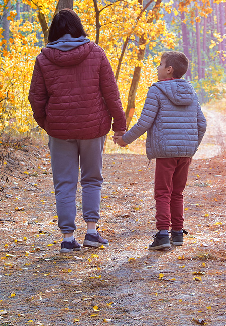 A mother and her young son hold hands as they stroll along a sunlit autumn forest path.