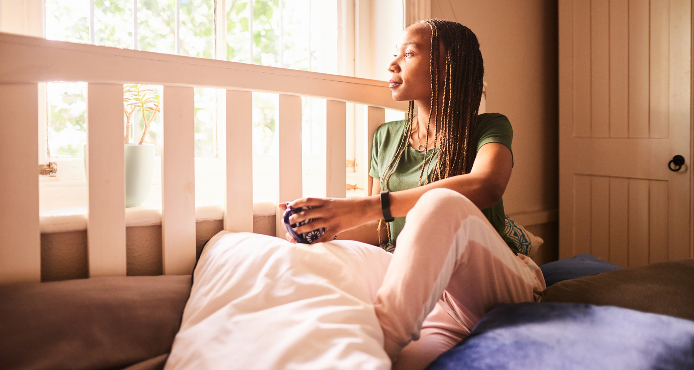 A person sitting on a bed looking out the window drinking coffee.