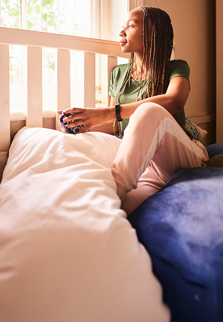 A person sitting on a bed looking out the window drinking coffee.