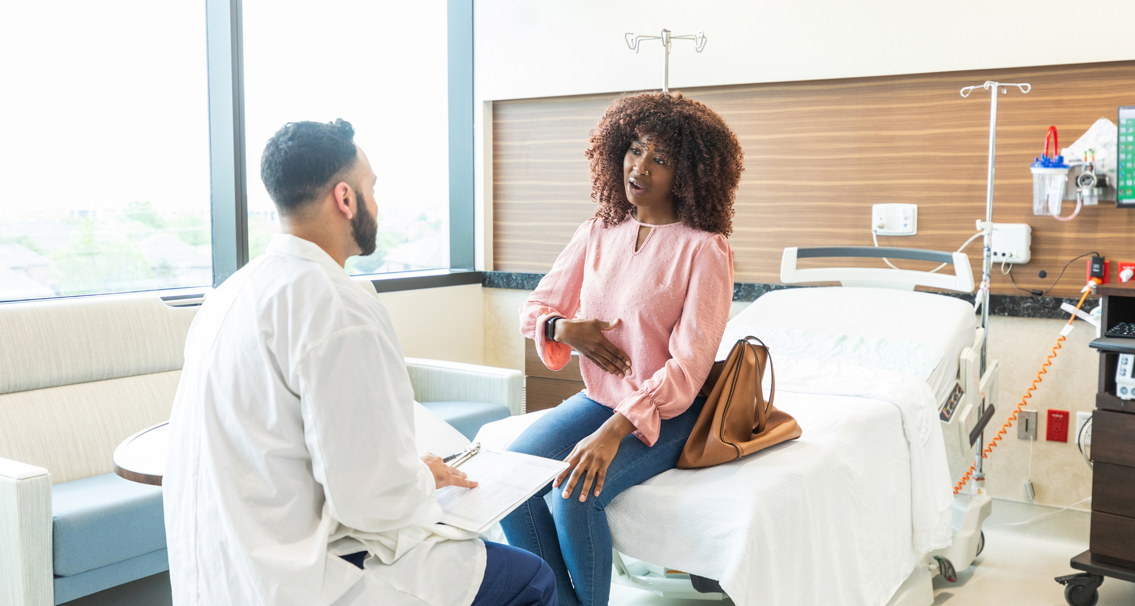 A woman sitting on a bed talking to a doctor about stomach pain.