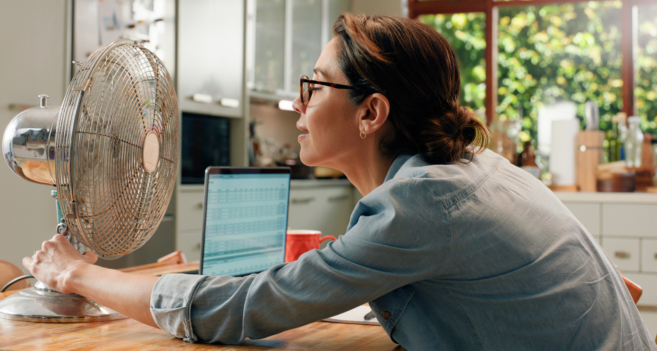 A woman experiencing relief from a fan during hot flashes at home while working.