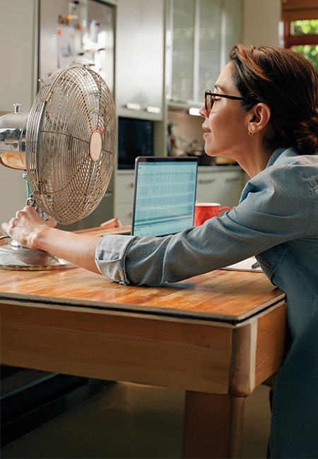 A woman experiencing relief from a fan during hot flashes at home while working.