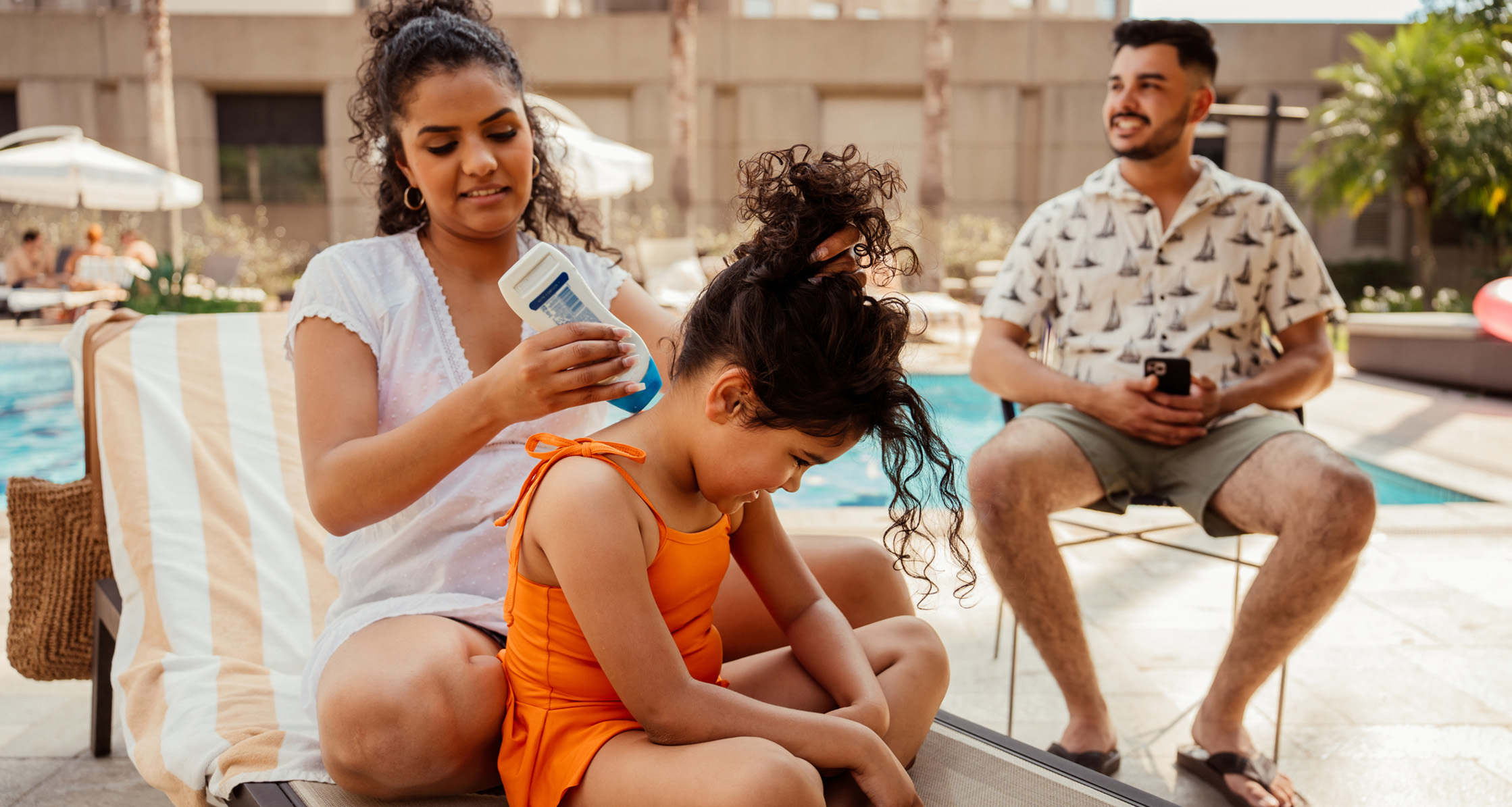 Mother applying sunscreen to daughter at the pool, with husband nearby.
