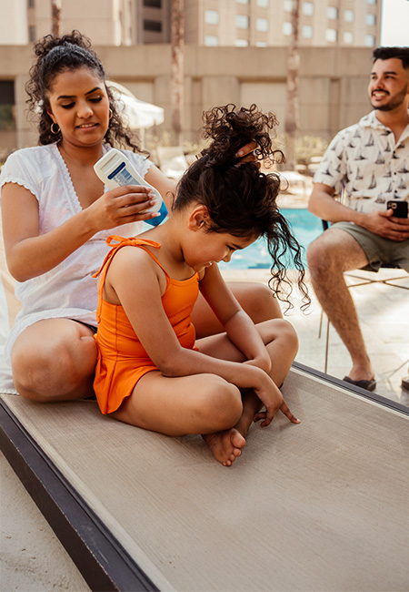 Mother applying sunscreen to daughter at the pool, with husband nearby.