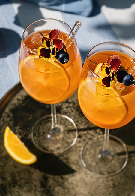 Two mocktail glasses of spritz with orange and flower garnish.