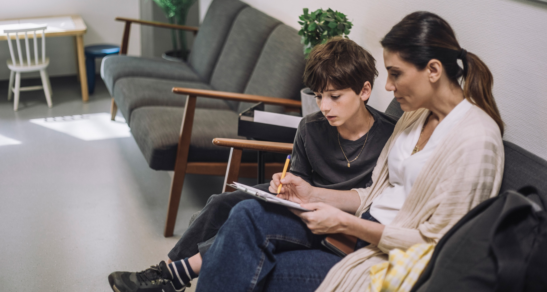 A mother with her son in urgent care waiting room, fills out paperwork.