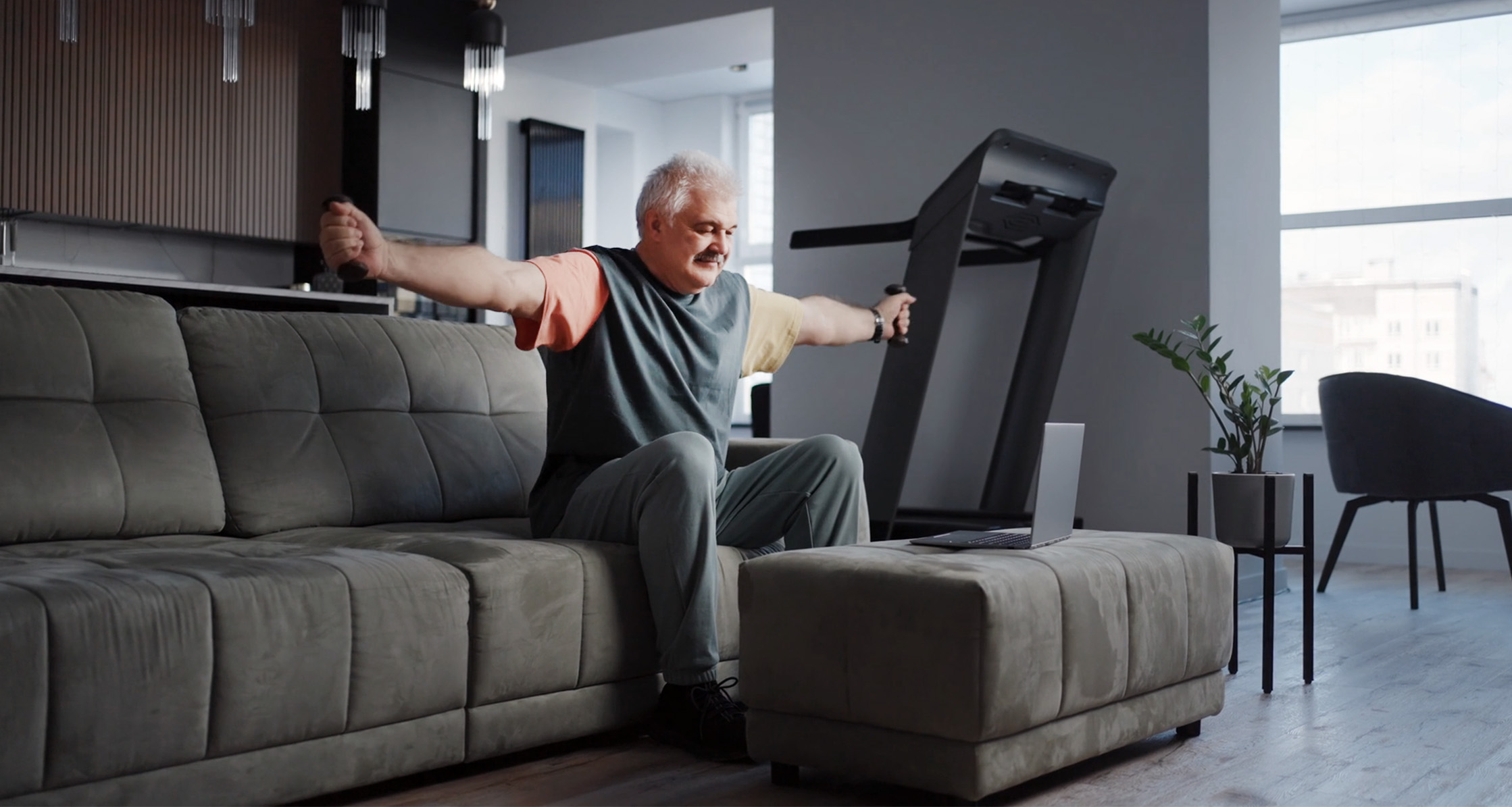 Person exercising at home seated on couch using light weights while following a workout on laptop.