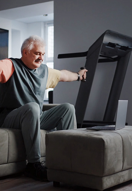 Person exercising at home seated on couch using light weights while following a workout on laptop.